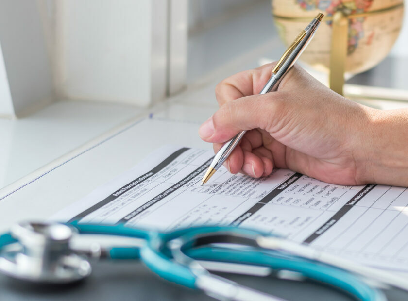 Doctor writing on medical health care record, patients discharge, or prescription form paperwork in hospital clinic office with physician's stethoscope on desk